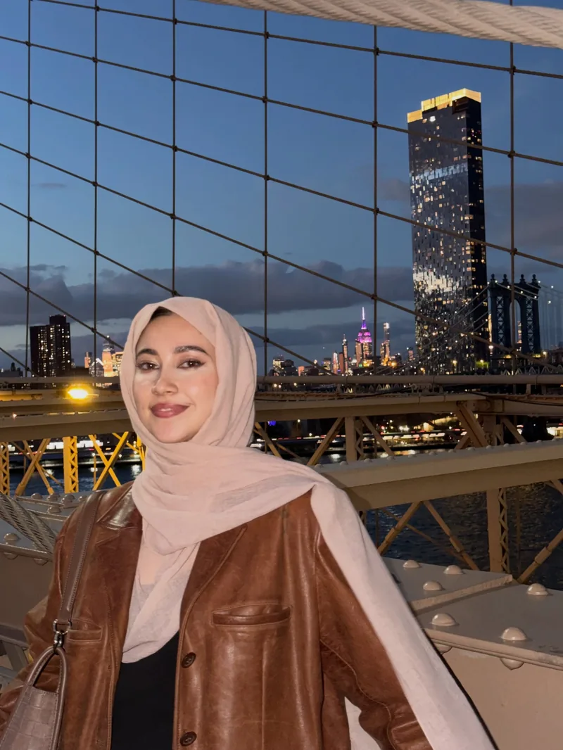Marifat Jamal smiling on the Brooklyn Bridge at dusk in a brown leather jacket, the Manhattan skyline lit behind her